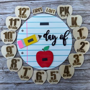 May include: A circular white board with blue horizontal lines and a pink vertical line resembling notebook paper. It features the black text "day of" and includes wooden apple-shaped cutouts labeled from Pre-K to 12th Grade, plus "FIRST" and "LAST" tags.