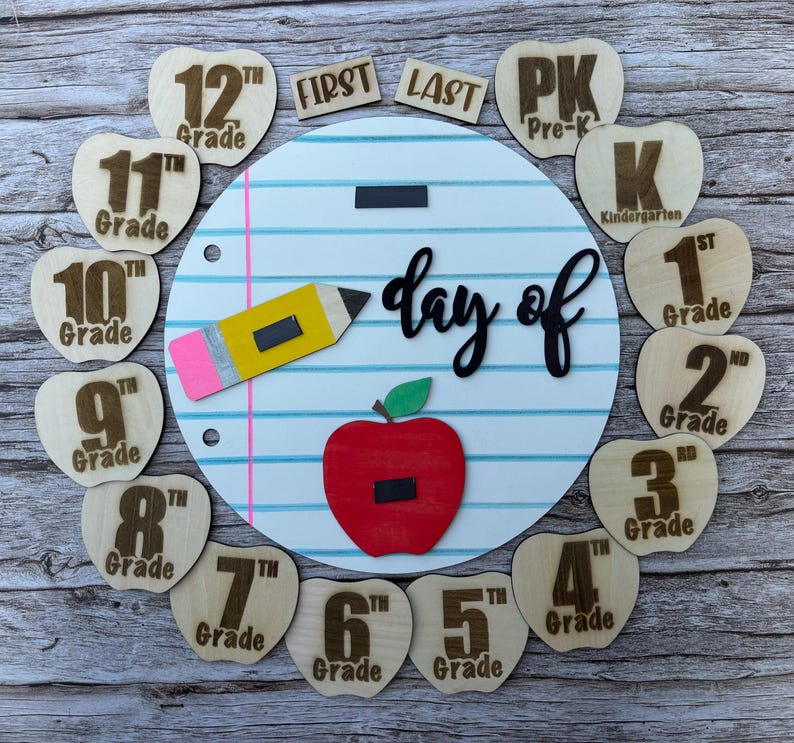 May include: A circular white board with blue horizontal lines and a pink vertical line resembling notebook paper. It features the black text "day of" and includes wooden apple-shaped cutouts labeled from Pre-K to 12th Grade, plus "FIRST" and "LAST" tags.