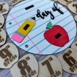 May include: A circular sign designed like notebook paper with the text day of, featuring a yellow pencil and a red apple. Surrounding the sign are smaller wooden apple-shaped cutouts engraved with various grade levels from PK to 12th grade.