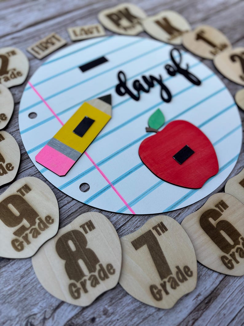 May include: A circular sign designed like notebook paper with the text day of, featuring a yellow pencil and a red apple. Surrounding the sign are smaller wooden apple-shaped cutouts engraved with various grade levels from PK to 12th grade.
