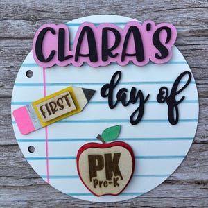 May include: A circular sign designed to look like notebook paper with blue horizontal lines and a pink vertical margin. It features the text CLARA'S day of in black and pink, a yellow pencil cutout labeled FIRST, and a red apple cutout labeled PK Pre-K.
