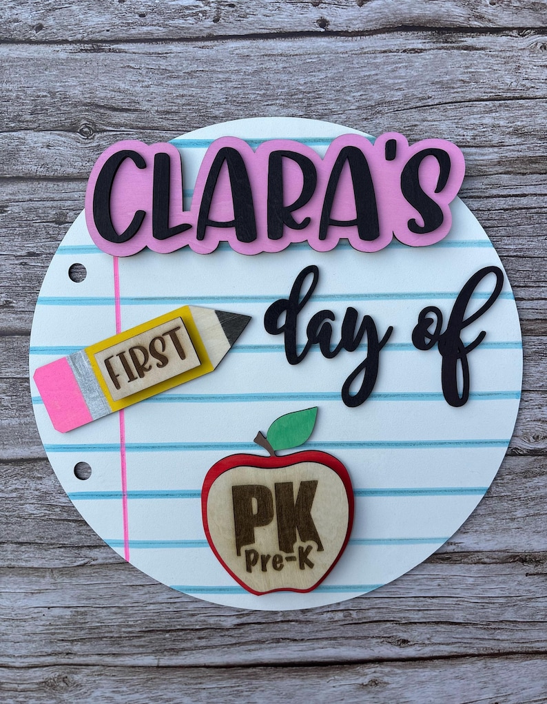 May include: A circular sign designed to look like notebook paper with blue horizontal lines and a pink vertical margin. It features the text CLARA'S day of in black and pink, a yellow pencil cutout labeled FIRST, and a red apple cutout labeled PK Pre-K.
