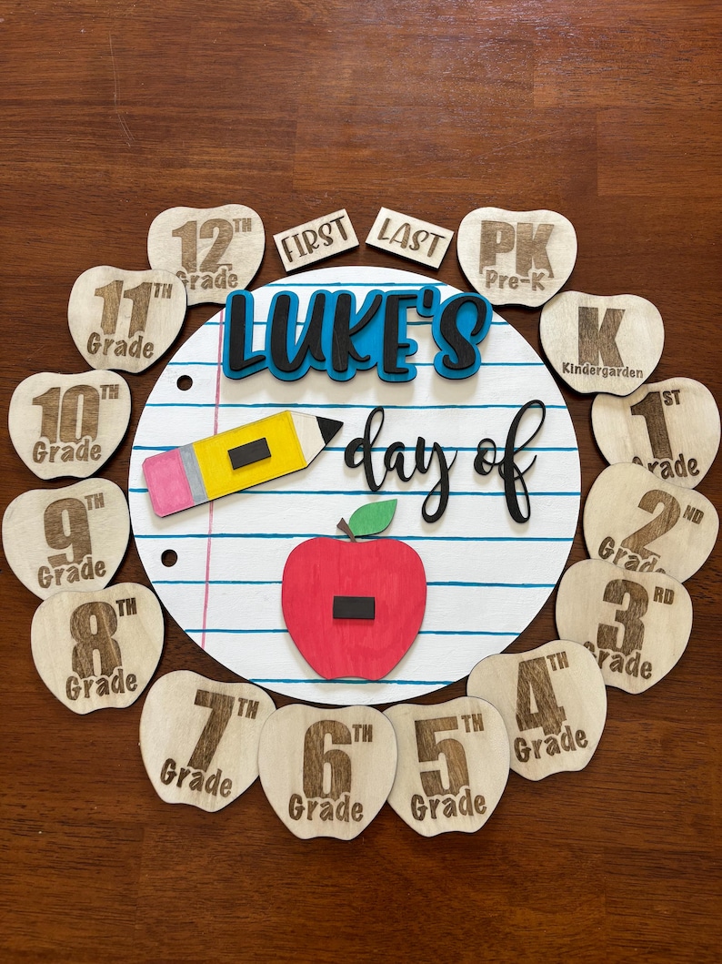 Op de afbeelding: Houten schoolbord met de tekst "Luke's day of" en cijfermarkeringen van Pre-K tot en met de 12e klas. Voorzien van een rode appel, een gele potlood en een notitiepapier ontwerp. Perfect voor back-to-school decoratie.