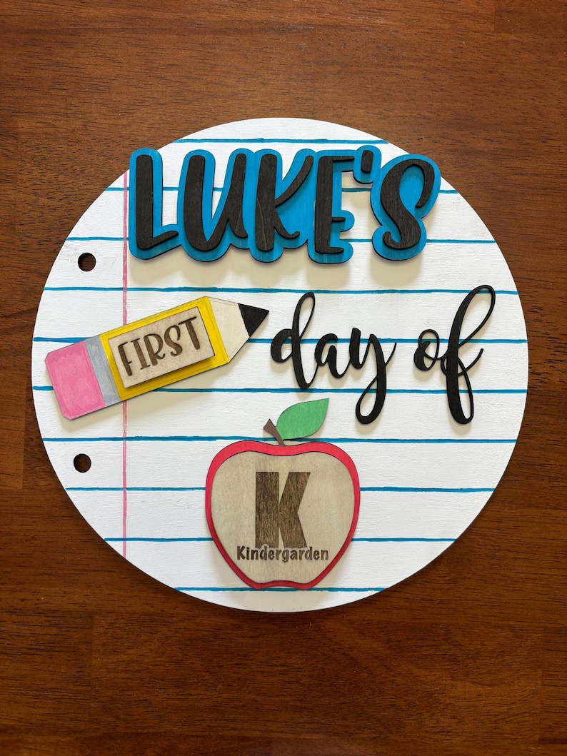 Op de afbeelding: Rond houten bord met een schoolthema. Het bord heeft blauwe lijnen, een potlood, een appel en de tekst "LUKE'S day of FIRST Kindergarten". Het bord staat op een bruin houten oppervlak.
