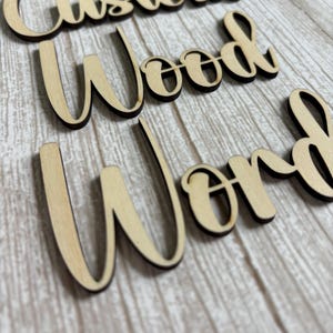 May include: Three separate words cut from light-colored wood with dark, charred edges. The words are arranged in a vertical stack and read Custom, Wood, and Word in a cursive script font. Each letter is connected to the next in a flowing style.