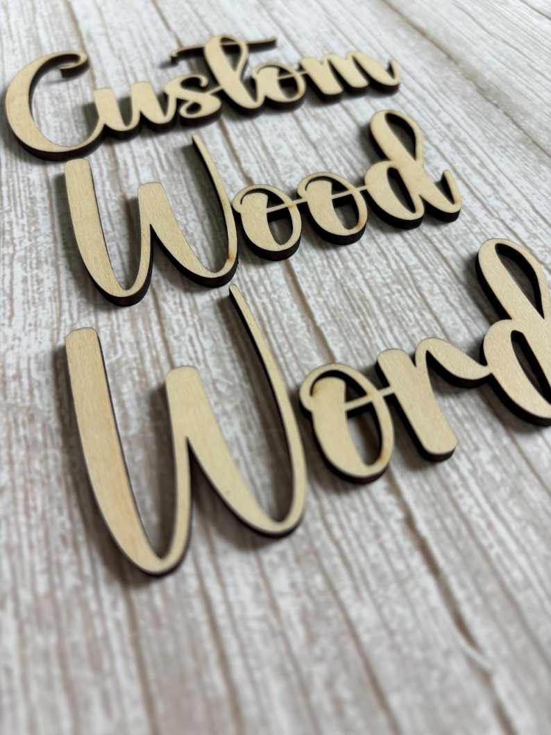 May include: Three separate words cut from light-colored wood with dark, charred edges. The words are arranged in a vertical stack and read Custom, Wood, and Word in a cursive script font. Each letter is connected to the next in a flowing style.