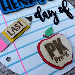 May include: A circular wooden sign designed to look like notebook paper with blue and pink lines. It features the text "HENRY'S last day of" in blue and black, a yellow pencil cutout with the word "LAST", and a red apple with "PK Pre-k".