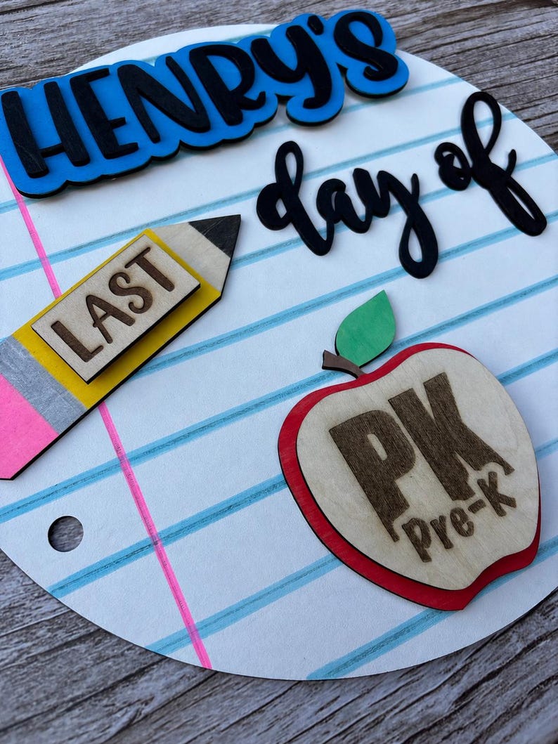 May include: A circular wooden sign designed to look like notebook paper with blue and pink lines. It features the text "HENRY'S last day of" in blue and black, a yellow pencil cutout with the word "LAST", and a red apple with "PK Pre-k".