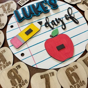 Op de afbeelding: Een houten schoolbord met de tekst "Luke's day of" en een potlood en appel. Inclusief houten appelvormige rangniveaumarkeringen en andere houten uitsparingen met tekst als "First", "Last" en "Pre-K".