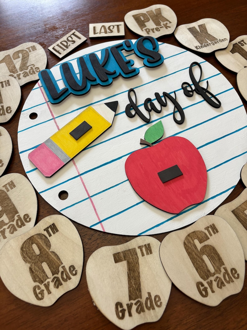 Op de afbeelding: Een houten schoolbord met de tekst "Luke's day of" en een potlood en appel. Inclusief houten appelvormige rangniveaumarkeringen en andere houten uitsparingen met tekst als "First", "Last" en "Pre-K".