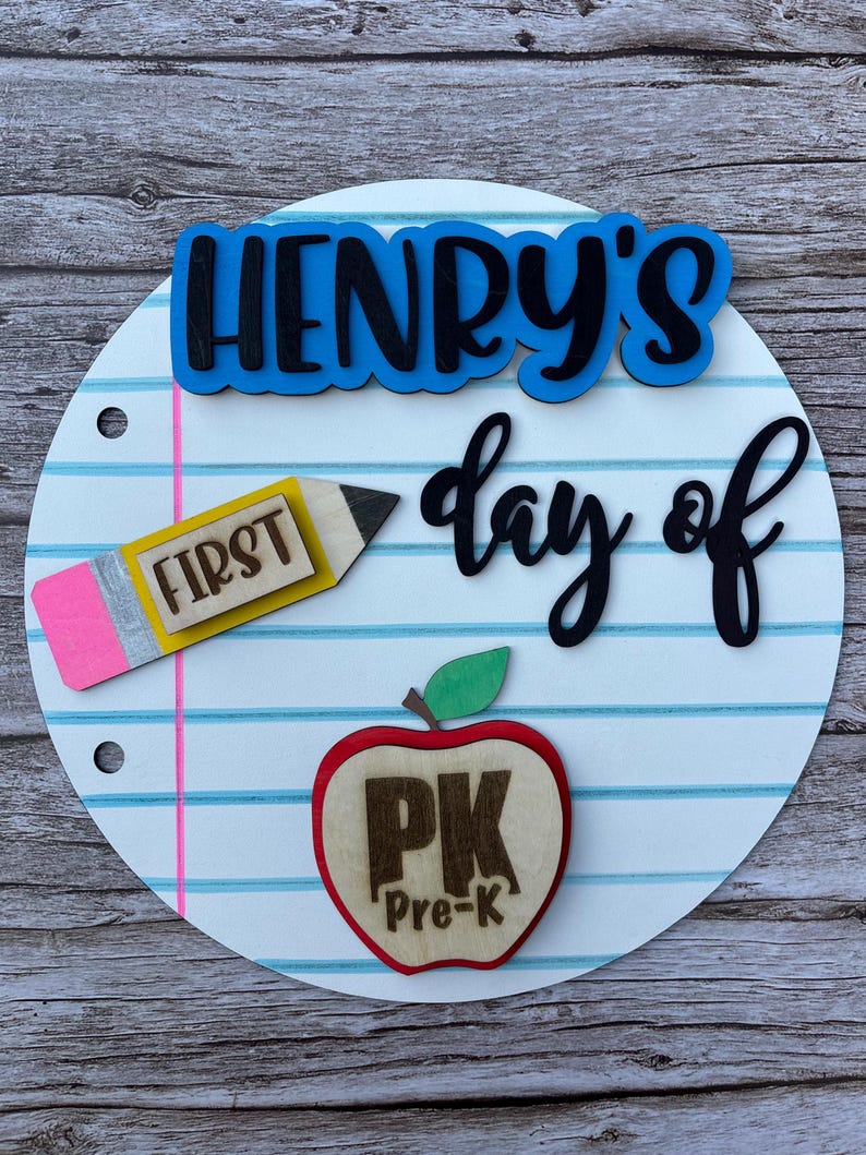May include: A circular sign designed to look like a piece of lined notebook paper with a pink margin line. It features the blue text "HENRY'S", the black text "day of", a yellow pencil cutout labeled "FIRST", and a red apple labeled "PK Pre-K".