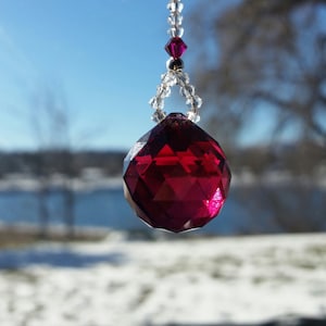 Op de afbeelding: Een gefacetteerd, dieprood kristallen ornament hangt aan een zilveren ketting met heldere kralen. Het kristal heeft een grote, ronde vorm met vele platte oppervlakken. De achtergrond is een wazig winterlandschap en blauwe lucht.