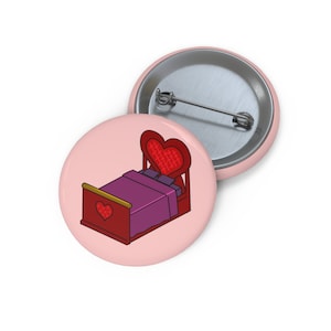 May include: A pink button featuring a cartoon illustration of a red bed with a heart-shaped headboard and a purple blanket.