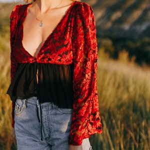 Puede incluir: Una blusa de terciopelo rojo con una superposición de gasa negra y un lazo en la parte delantera. La blusa tiene un escote en V profundo y mangas largas.