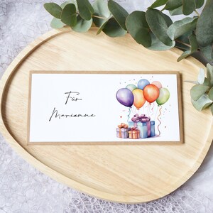 Könnte beinhalten: Eine Grußkarte mit dem Text "Für Marianne" und einer Aquarellillustration von bunten Luftballons und verpackten Geschenken. Die Karte liegt auf einem Holztablett mit Eukalyptusblättern.