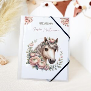 Könnte beinhalten: Ein transparenter Kunststoffordner mit einer Aquarellillustration eines Pferdekopfes, verziert mit einem Blumenkranz und einem Kranz aus rosa Rosen. Auf dem Cover steht "MEINE SAMMELMAPPE" und "Sophia Mustermann". Ein schwarzes Gummiband sichert den Ordner.