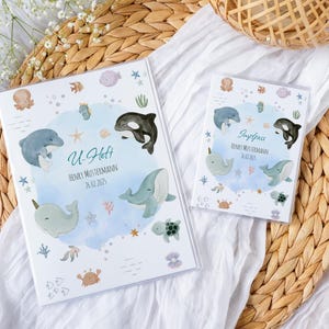 Könnte beinhalten: Zwei weiße Bücher mit aquatischen Illustrationen. Das größere Buch trägt den Text "U-Heft" und den Namen "Henry Mustermann" mit einem Datum. Das kleinere Buch trägt den Text "Impfpass" und denselben Namen und dasselbe Datum. Die Illustrationen zeigen Wale, Delfine und andere Meeresbewohner.