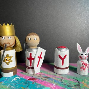 May include: Wooden peg dolls depicting a king, knight, and two other figures. The king wears a gold crown and has a sword. The knight has a shield with a red cross. The other figures have red and white designs.