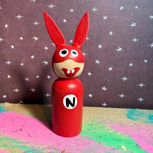 May include: A red wooden peg doll in the shape of a rabbit with large ears. The doll has a white face with black eyes and a red mask. A white letter "N" is on the doll's red body. The background is a dark purple with white stars.