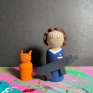 May include: Wooden peg dolls: an orange cat with white eyes and a pink nose, a person in a blue uniform holding a gray gun. The cat is next to the person. The background is a mix of colors.