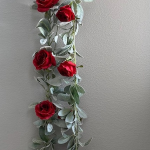 May include: A decorative garland featuring vibrant red roses and silvery-green leaves. The floral arrangement is hanging against a neutral-colored wall, creating a visually appealing contrast. This garland is perfect for adding a touch of elegance to any space.