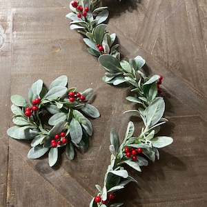 May include: A garland of faux lambs ear greenery with red berries. The garland is arranged in a decorative pattern on a wooden surface.