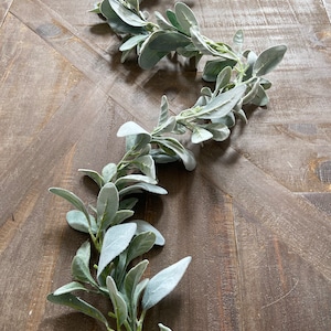 May include: A long garland of faux lambs ear leaves, with a light green color and a fuzzy texture, is displayed on a wooden surface.