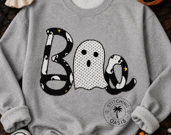 May include: A heather gray crewneck sweatshirt featuring a large graphic on the front that spells the word Boo. The letter B and the letter o are black with white ghost patterns and small yellow stars, while the middle letter o is a white ghost.