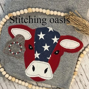 May include: A gray t-shirt with a red, white, and blue cow applique. The cow has a star-spangled design on its head and a red and white tag on its ear. The text "Stitching oasis" is visible above the cow.