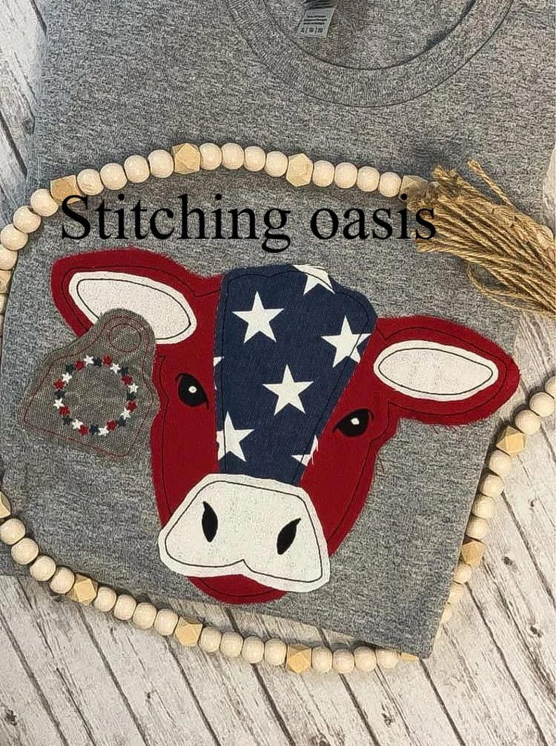 May include: A gray t-shirt with a red, white, and blue cow applique. The cow has a star-spangled design on its head and a red and white tag on its ear. The text "Stitching oasis" is visible above the cow.