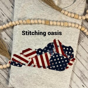 Puede incluir: Una camiseta gris con el texto "Stitching oasis" impreso en la parte delantera. La camiseta presenta un dise&ntilde;o de bandera estadounidense en forma del estado de Kentucky.