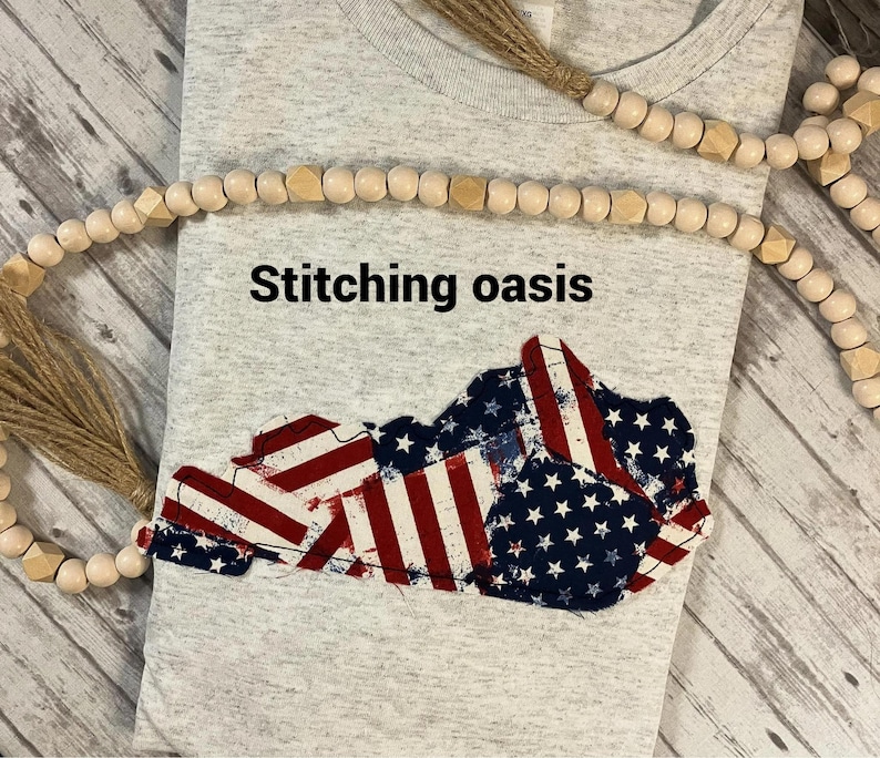 Puede incluir: Una camiseta gris con el texto "Stitching oasis" impreso en la parte delantera. La camiseta presenta un dise&ntilde;o de bandera estadounidense en forma del estado de Kentucky.