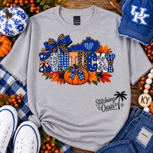May include: A heather gray t-shirt featuring a graphic of the word KENTUCKY in blue and white patterns, a pumpkin, a blue bow, and autumn leaves. The text Stitching Oasis with a palm tree icon is printed in black on the lower right side.