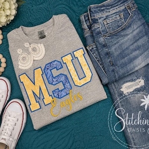 May include: A heather gray crewneck sweatshirt featuring the letters MSU in yellow and blue paisley print, with the word Eagles in yellow script below. The shirt is paired with blue denim jeans, white canvas sneakers, and white beaded earrings.