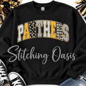 May include: A black crewneck sweatshirt featuring the word PANTHERS in an arched, block-letter font. The letters have varied patterns including black and white polka dots, solid gold, black and white buffalo plaid, and silver glitter textures.