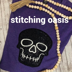 May include: Purple t-shirt with a black skull embroidered on the front. The skull has white stitching and rhinestones. The shirt is decorated with a wooden bead garland with tassels.