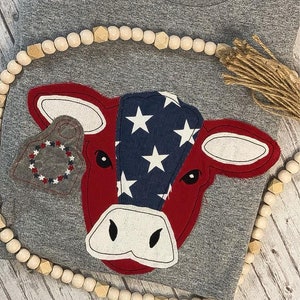 May include: A gray t-shirt with a red and blue cow applique. The cow has a white nose and eyes and is decorated with white stars on a blue background. The cow is wearing a gray tag with red and white trim.