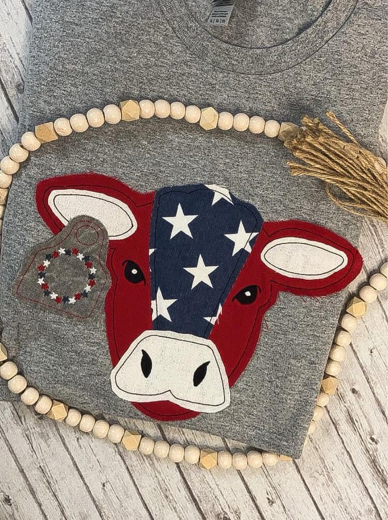 May include: A gray t-shirt with a red and blue cow applique. The cow has a white nose and eyes and is decorated with white stars on a blue background. The cow is wearing a gray tag with red and white trim.