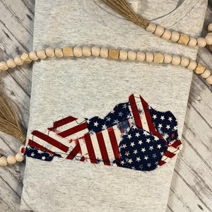 Puede incluir: Una camiseta gris con un dise&ntilde;o de bandera estadounidense desgastado en forma del estado de Kentucky.
