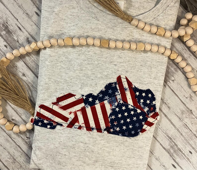Puede incluir: Una camiseta gris con un dise&ntilde;o de bandera estadounidense desgastado en forma del estado de Kentucky.