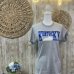 May include: Gray t-shirt with a blue and white Kentucky state outline and the word "Kentucky" in blue sequins.