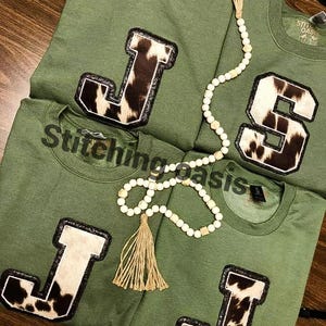 May include: Three green t-shirts with cowhide patches in the shape of the letter J. The shirts are stacked on top of each other. A white beaded necklace with a brown tassel hangs over the shirts.