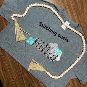 May include: A gray t-shirt with a black and white design of the state of Kentucky. The shirt has the text "Stitching Oasis" printed on it. A wooden bead garland with tassels is draped over the shirt.