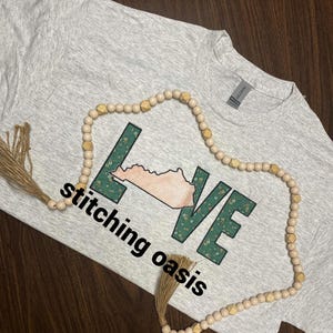 May include: A gray t-shirt with the word "LOVE" spelled out in green with a floral pattern. The letter "O" is replaced with the outline of the state of Kentucky. The text "stitching oasis" is printed below the word "LOVE". A wooden beaded necklace with a tassel is draped over the shirt.