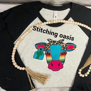 May include: A black and grey baseball tee shirt with a colorful embroidered cow wearing a bandana. The shirt has the text "Stitching Oasis" on the front. The shirt is decorated with a wooden bead necklace with a tassel.