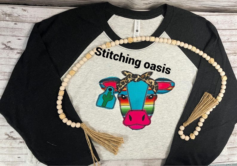 May include: A black and grey baseball tee shirt with a colorful embroidered cow wearing a bandana. The shirt has the text "Stitching Oasis" on the front. The shirt is decorated with a wooden bead necklace with a tassel.
