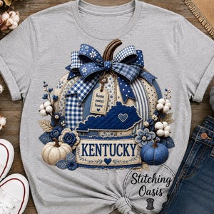 May include: A heather gray t-shirt featuring a large graphic of a pumpkin decorated with blue and white patterns, a plaid bow, cotton stems, and a tag that reads home sweet home. The design includes a blue Kentucky state outline and the word KENTUCKY.
