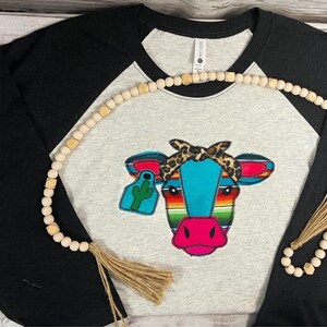 May include: A black and grey baseball tee shirt with a colorful cow design. The cow is wearing a leopard print bandana and has a green cactus on its ear tag. The shirt is decorated with a wooden bead necklace with a tassel.