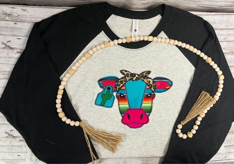 May include: A black and grey baseball tee shirt with a colorful cow design. The cow is wearing a leopard print bandana and has a green cactus on its ear tag. The shirt is decorated with a wooden bead necklace with a tassel.
