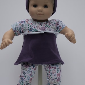 May include: A doll wearing a purple and white floral print outfit. The doll has blonde hair and blue eyes. The outfit includes a short-sleeved top, leggings, and a headband.
